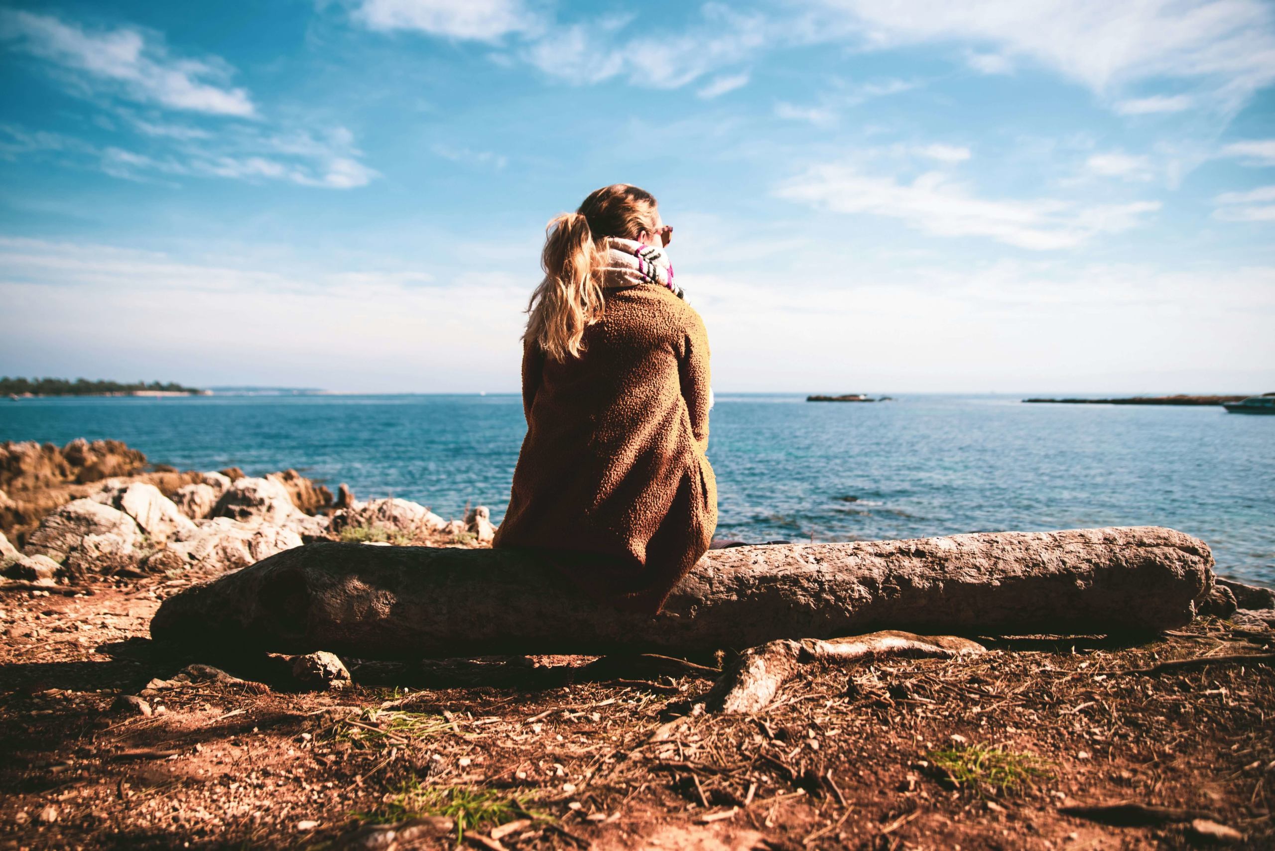 Image of a woman sitting on a log overlooking water on a sunny day. Discover how empath counseling in Austin, TX can help you with your emotions.